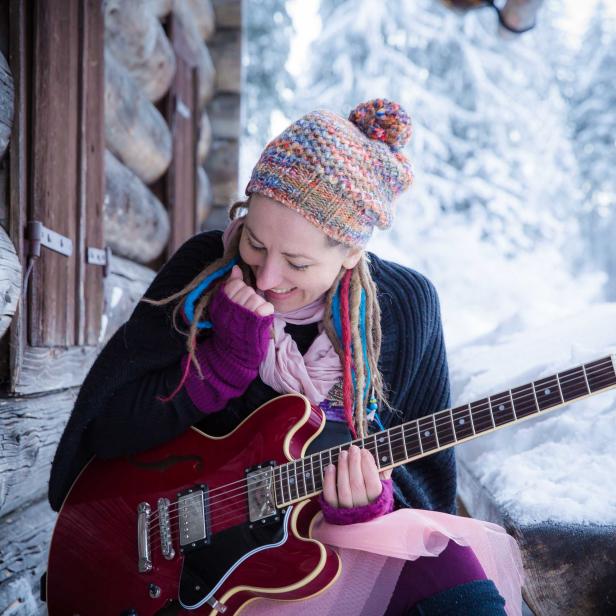Eine Frau mit Mütze und Gitarre sitzt vor einer verschneiten Holzhütte.