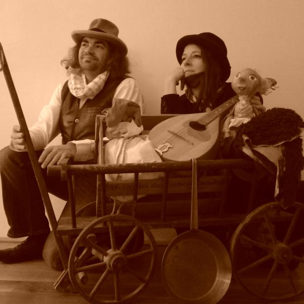Ein Mann und eine Frau sitzen mit einer Mandoline und einer Puppe in einem Leiterwagen.