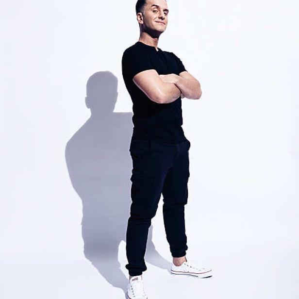 Ein Mann mit verschränkten Armen, bekleidet mit einem schwarzen T-Shirt und dunkler Hose, steht vor einem weißen Hintergrund.