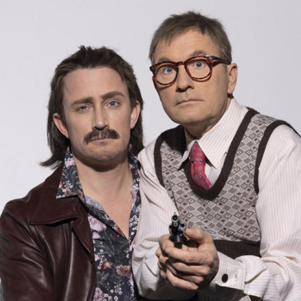 Zwei Männer posieren, einer mit Schnurrbart und Lederjacke, der andere mit Weste und Pistole.