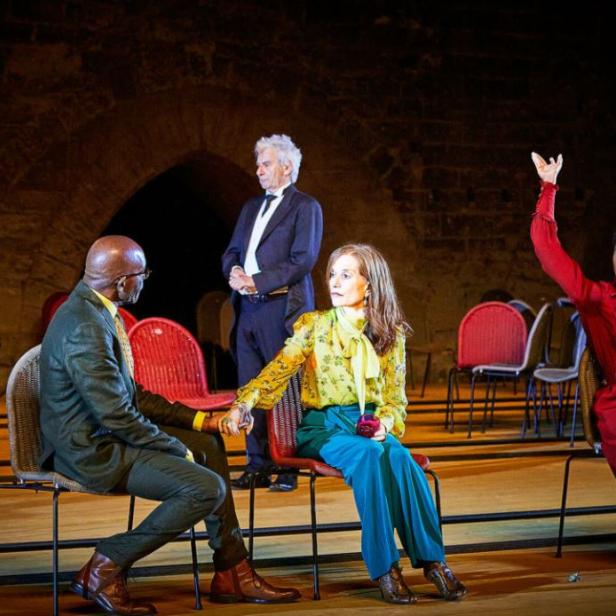 Szene aus einer Theateraufführung mit mehreren Darstellern auf Stühlen.