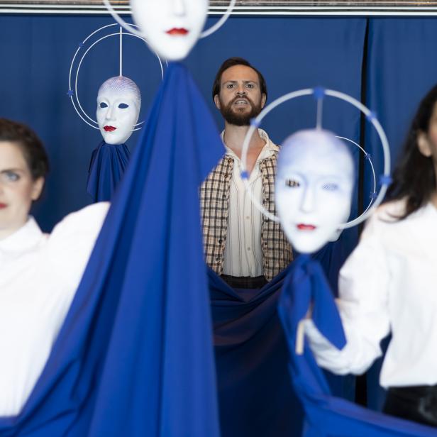 Eine Performance mit drei Personen und schwebenden, weißen Masken vor blauem Hintergrund.