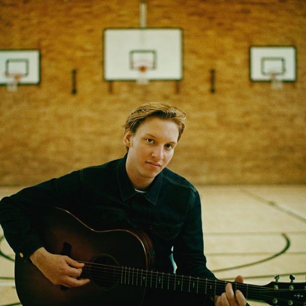 Ein Mann mit blonder Haaren spielt Gitarre in einer Turnhalle.