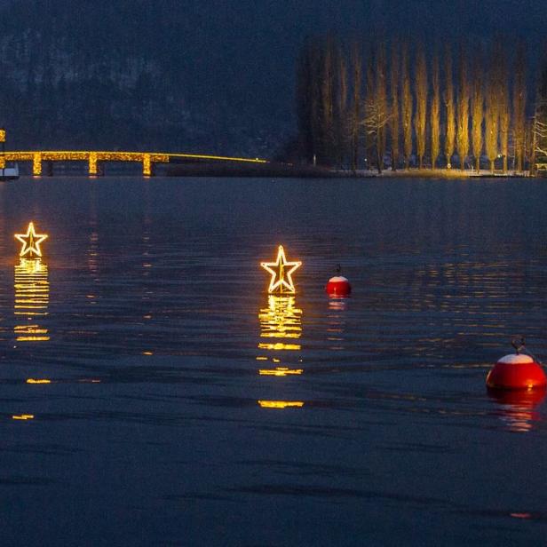 Mehrere beleuchtete Sterne schwimmen auf einem See, im Hintergrund ein beleuchtetes Gebäude.
