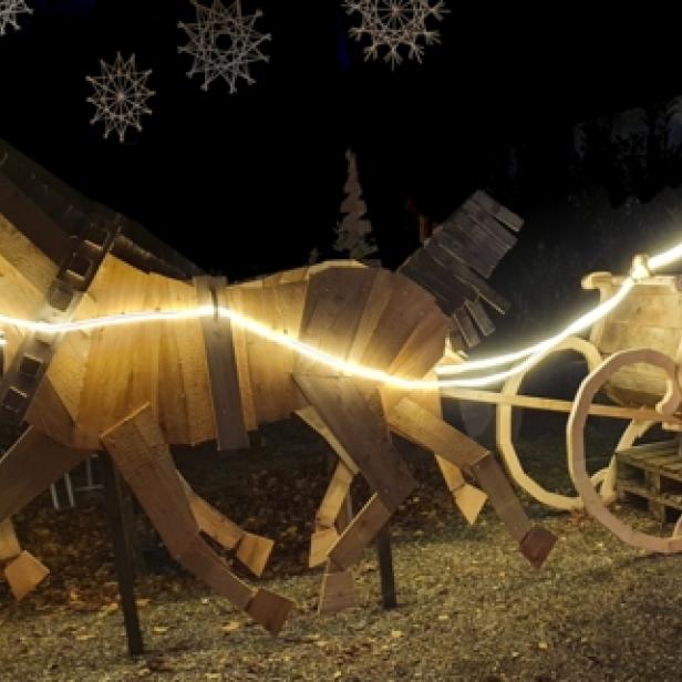 Ein Mann sitzt auf einem Holzschlitten, der von zwei hölzernen Pferden gezogen wird, alles mit Lichtern geschmückt.