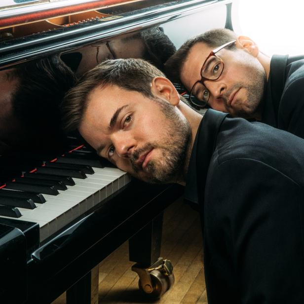 Zwei Männer in Anzügen liegen an einem Klavier.