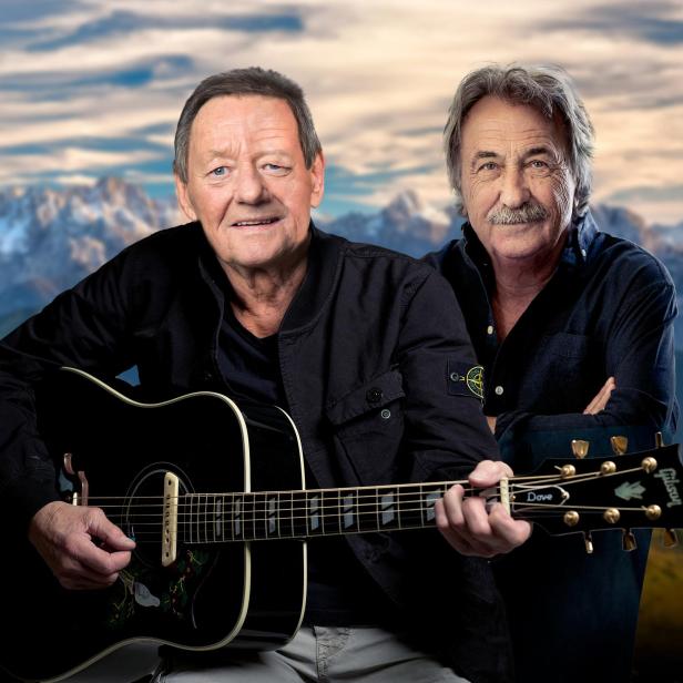 Zwei Männer posieren mit einer Gitarre vor einem Bergpanorama.