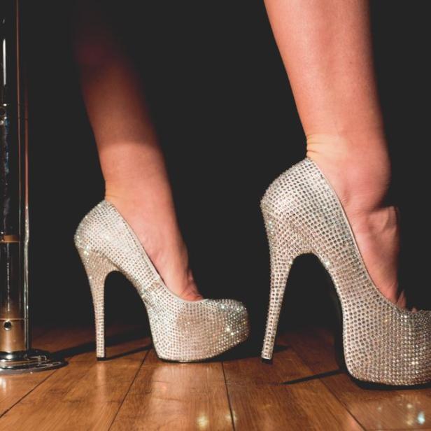 Nahaufnahme von Füßen in silbernen, mit Strass besetzten High Heels neben einer Stange.