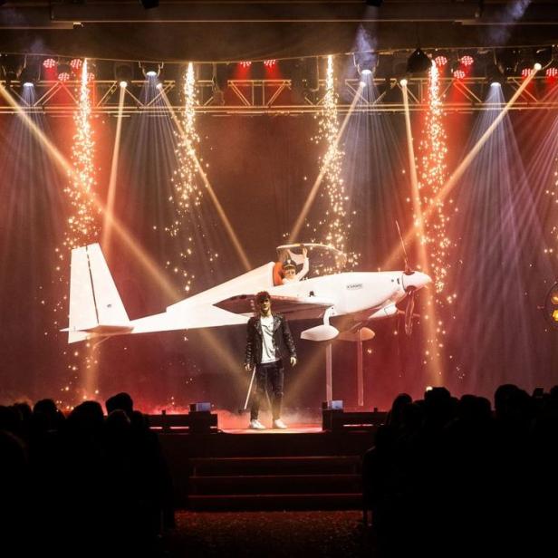 Ein Mann steht vor einem kleinen Flugzeug auf einer Bühne mit Lichteffekten und Publikum.