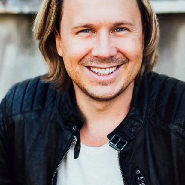 Ein lächelnder Mann mit blonden Haaren und Lederjacke vor einer grauen Wand.