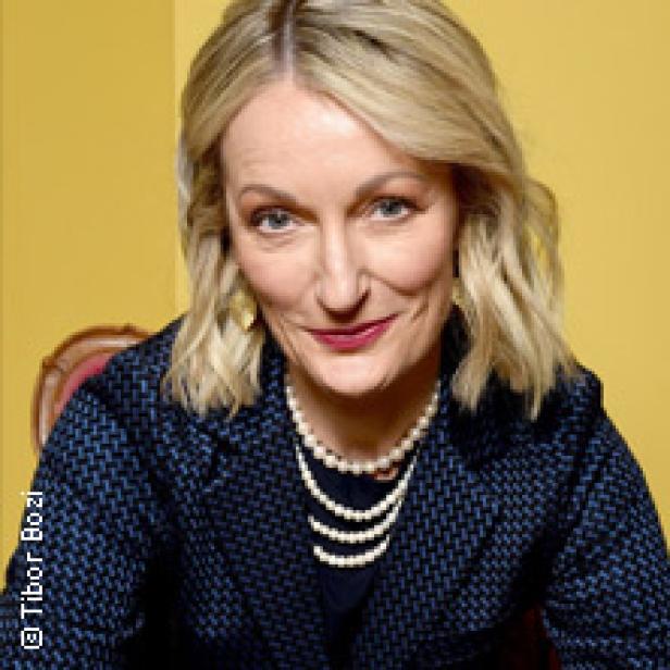 Eine blonde Frau mit Perlenkette lächelt in die Kamera.