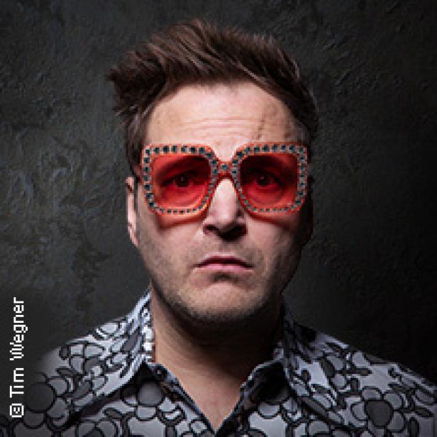 Ein Mann mit auffälliger Brille und gemustertem Hemd blickt in die Kamera.