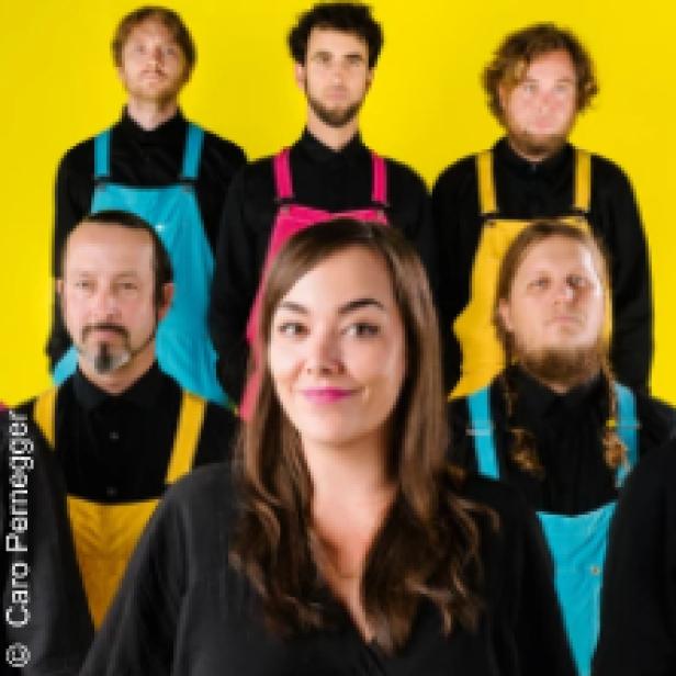 Eine Musikgruppe mit sechs Mitgliedern posiert vor einem gelben Hintergrund.