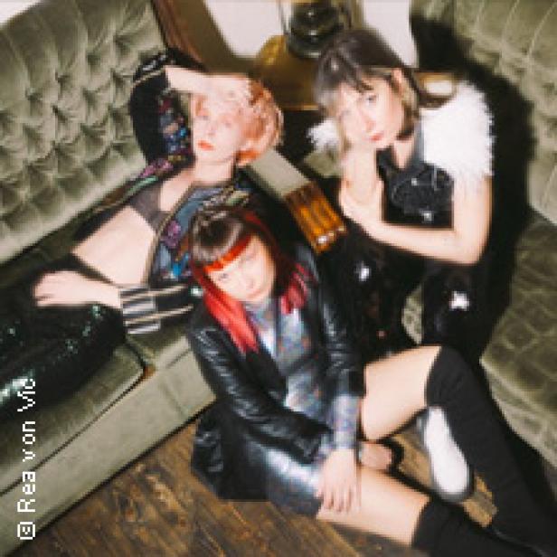 Drei Frauen posieren in glitzernden Outfits auf einem grünen Sofa.