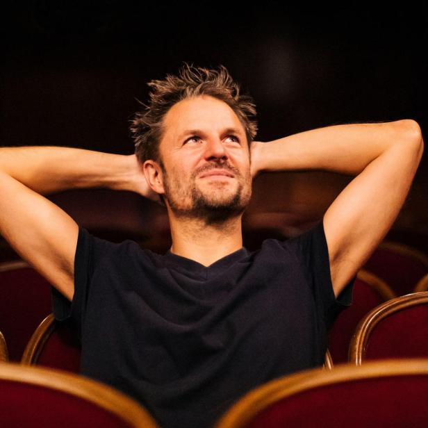 Philipp Hochmair sitzt entspannt mit verschränkten Armen in einem Theatersaal.