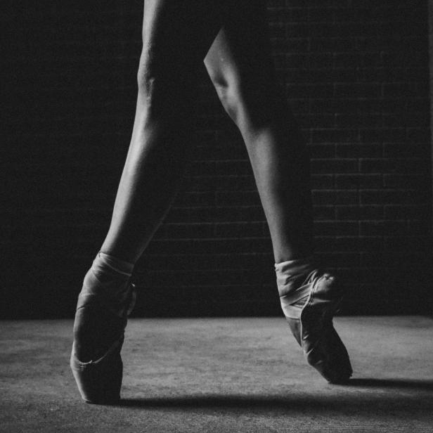 Die Füße einer Ballerina in Spitzenschuhen vor einer dunklen Ziegelwand.
