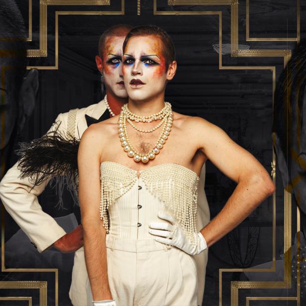 Zwei Dragqueens posieren vor einem dunklen Hintergrund mit dem Schriftzug „Hotel is gay“.