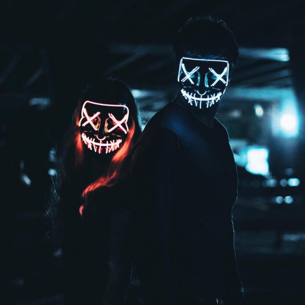 Zwei Personen tragen leuchtende Masken mit einem „X“ für die Augen und einem grinsenden Mund.
