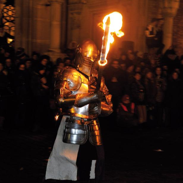 Ein Ritter in Rüstung hält eine brennende Fackel in der Hand, umgeben von einer Menschenmenge.
