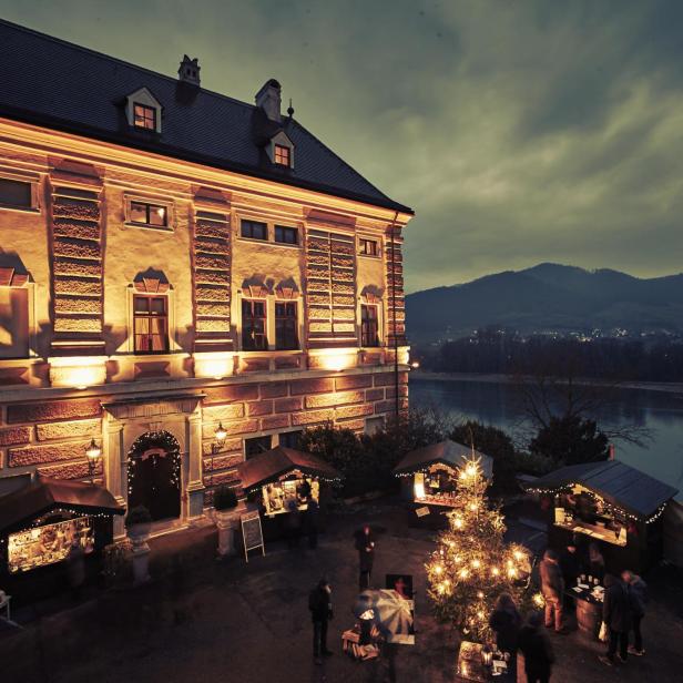 Ein Weihnachtsmarkt vor einem beleuchteten Gebäude mit Blick auf einen Fluss.