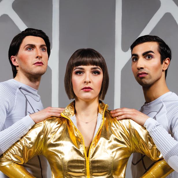 Eine Frau in goldener Jacke posiert mit zwei Männern in futuristischer Kleidung.