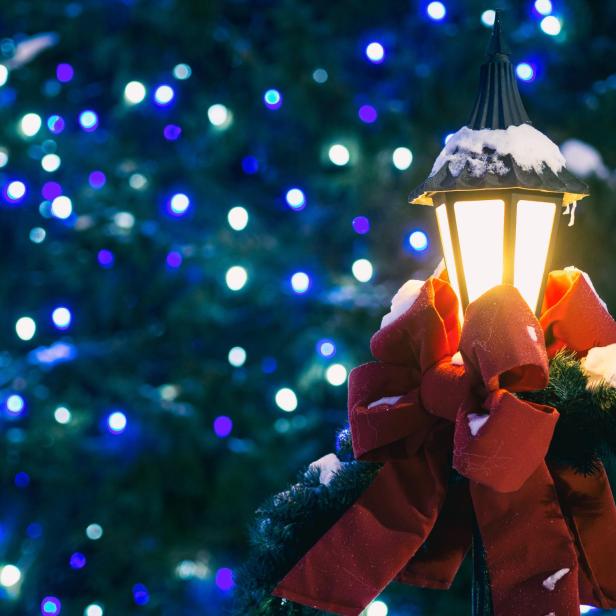 Eine schneebedeckte Laterne mit einer roten Schleife vor einem Weihnachtsbaum mit Lichtern.