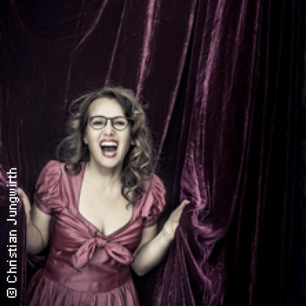 Eine Frau mit Brille und rotem Kleid steht vor einem roten Vorhang und lacht.