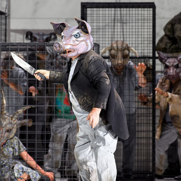 Ein Mann mit Schweinsmaske hält ein Messer, während andere Personen mit Tiermasken in Käfigen gefangen sind.