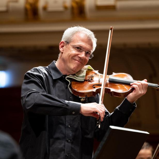 Ein Mann mit Brille spielt Geige in einem Orchester.