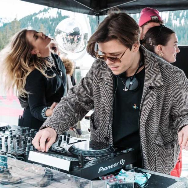Ein DJ legt auf einer Party im Freien Musik auf, umgeben von feiernden Menschen.