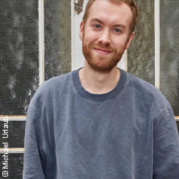 Ein junger Mann mit Bart und blauem Pullover lächelt in die Kamera.
