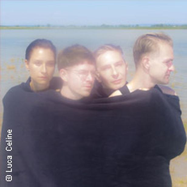 Vier Personen stehen am Wasser und sind in ein schwarzes Tuch gehüllt.