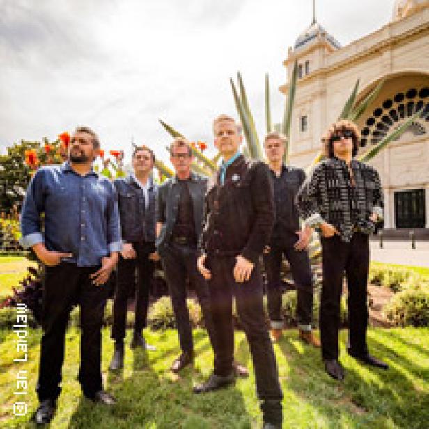 Eine Band posiert vor einem Gebäude mit Säulen und einem grünen Park im Vordergrund.