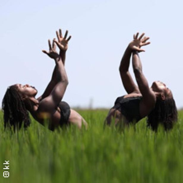 Zwei Frauen mit Dreadlocks tanzen in einem grünen Feld mit erhobenen Händen.