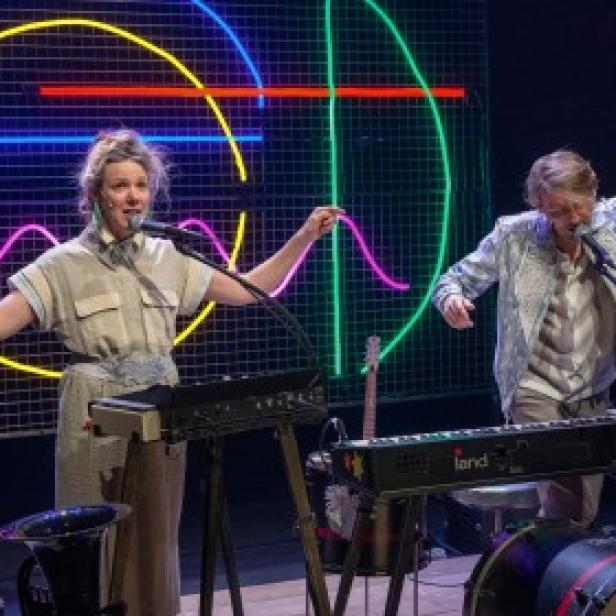 Zwei Musiker treten mit Keyboards und Mikrofonen vor einem neonbeleuchteten Hintergrund auf.