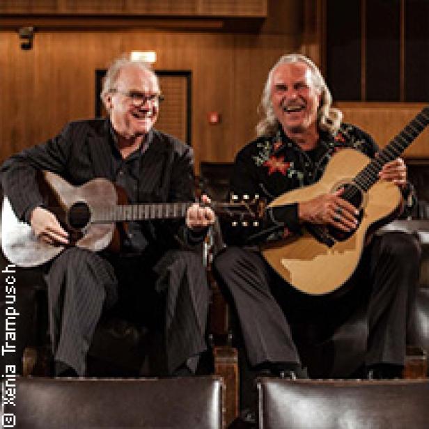 Zwei Männer sitzen mit Gitarren in einem Konzertsaal und lachen.