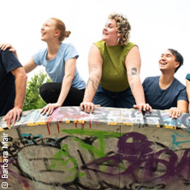 Eine Gruppe junger Leute steht an einer mit Graffiti bemalten Mauer und schaut nach oben.