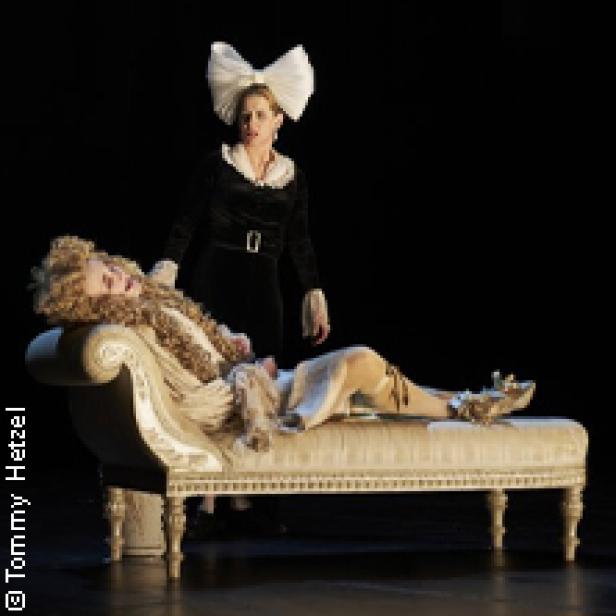 Eine Frau mit Schleife im Haar steht neben einer auf einer Couch liegenden Person.