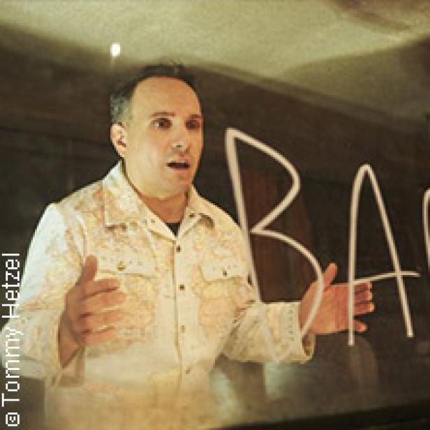 Ein Mann mit einem Hemd mit Weltkartenmuster gestikuliert vor einer Tafel mit der Aufschrift „BAR“.