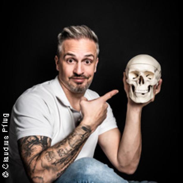 Ein Mann mit Tätowierungen zeigt auf einen menschlichen Schädel.