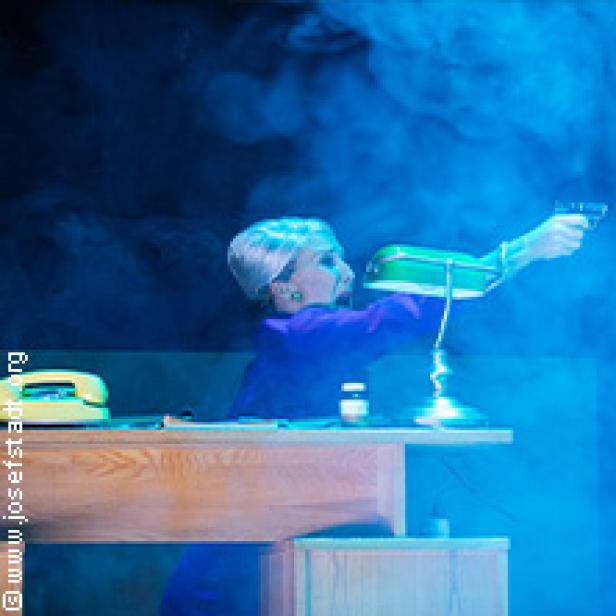 Eine Frau mit weißem Haar zielt mit einer Pistole über einen Schreibtisch.