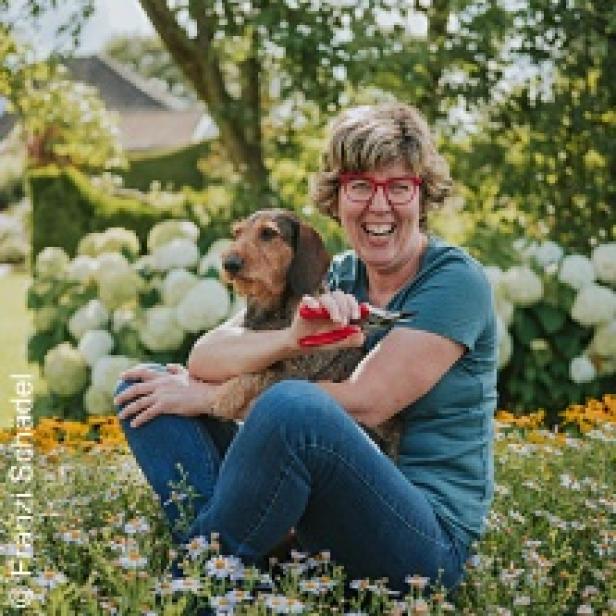 Eine Frau mit roter Brille sitzt mit einem Dackel auf einer Blumenwiese und hält eine Gartenschere.