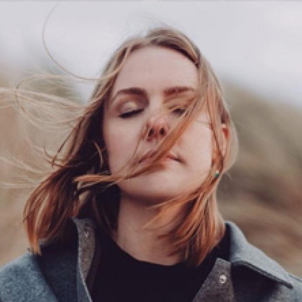 Eine junge Frau mit blonden Haaren und geschlossenen Augen steht im Wind.