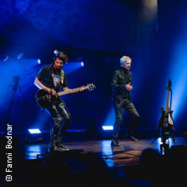 Zwei Musiker stehen auf einer Bühne, einer spielt Gitarre, der andere singt.