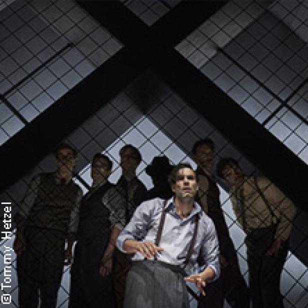 Eine Bühnenszene mit einem Mann im Vordergrund und mehreren Männern im Hintergrund vor einem Gitter.