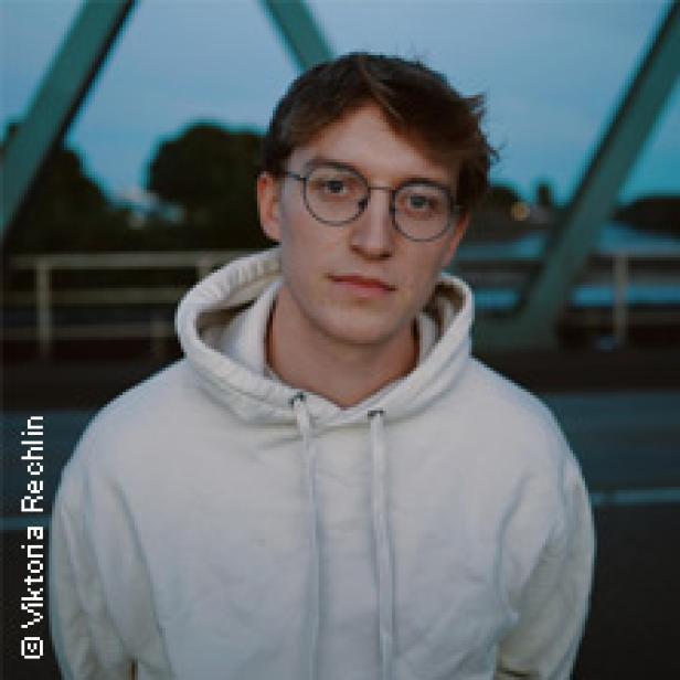 Ein junger Mann mit Brille und hellem Hoodie steht vor einer Brücke.