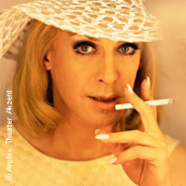 Ein Mann mit blonden Haaren, Hut und Zigarette blickt in die Kamera.