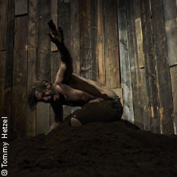 Ein Mann, bedeckt mit Schmutz, kauert auf einem Erdhaufen vor einer Holzwand.