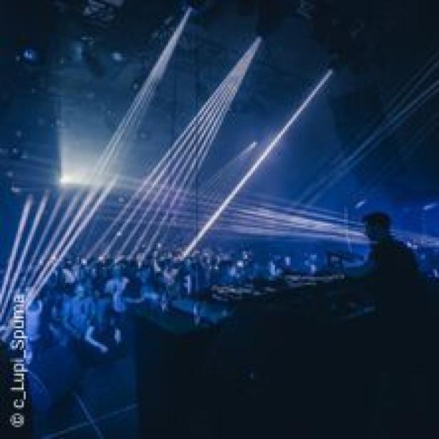 Ein DJ legt in einem dunklen Club vor einer tanzenden Menschenmenge auf, während Laserstrahlen durch den Raum schneiden.