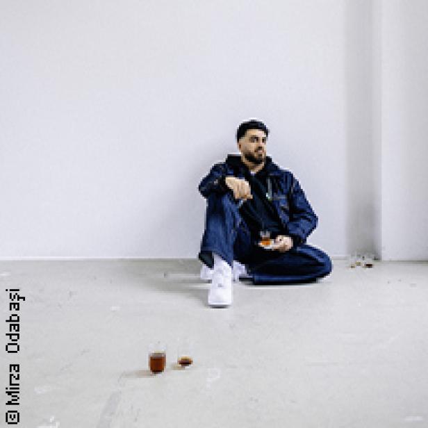 Ein Mann sitzt auf dem Boden in einem leeren Raum und hält ein Glas in der Hand.